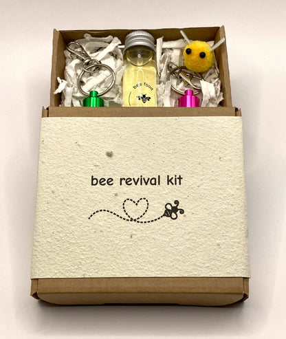 Showing bee revival kit box, slightly opened showing the contents, consisting of glass bottle with 20ml Inveråtbee syrup, two bee revival keyrings, seed paper packing and a cute little fabric bee.